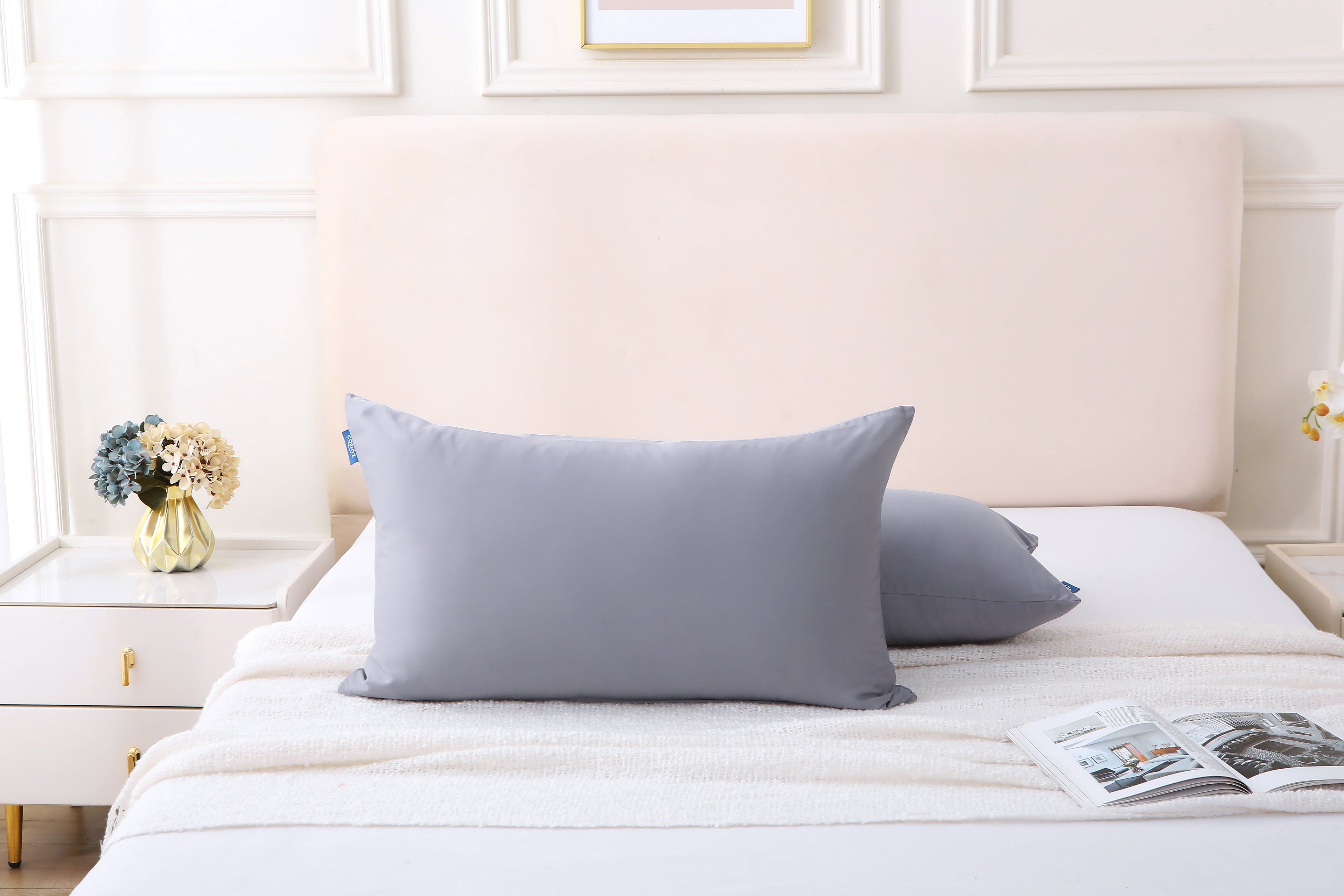 Cushyz gray pillows arranged on a bed in a cozy bedroom setting with a decorative vase and a magazine on the side.